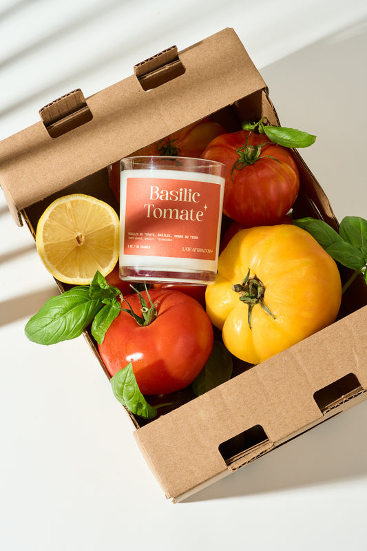 Cardboard box with tomatoes, a lemon, and a candle labeled 'Basilic Tomate' on a white background.