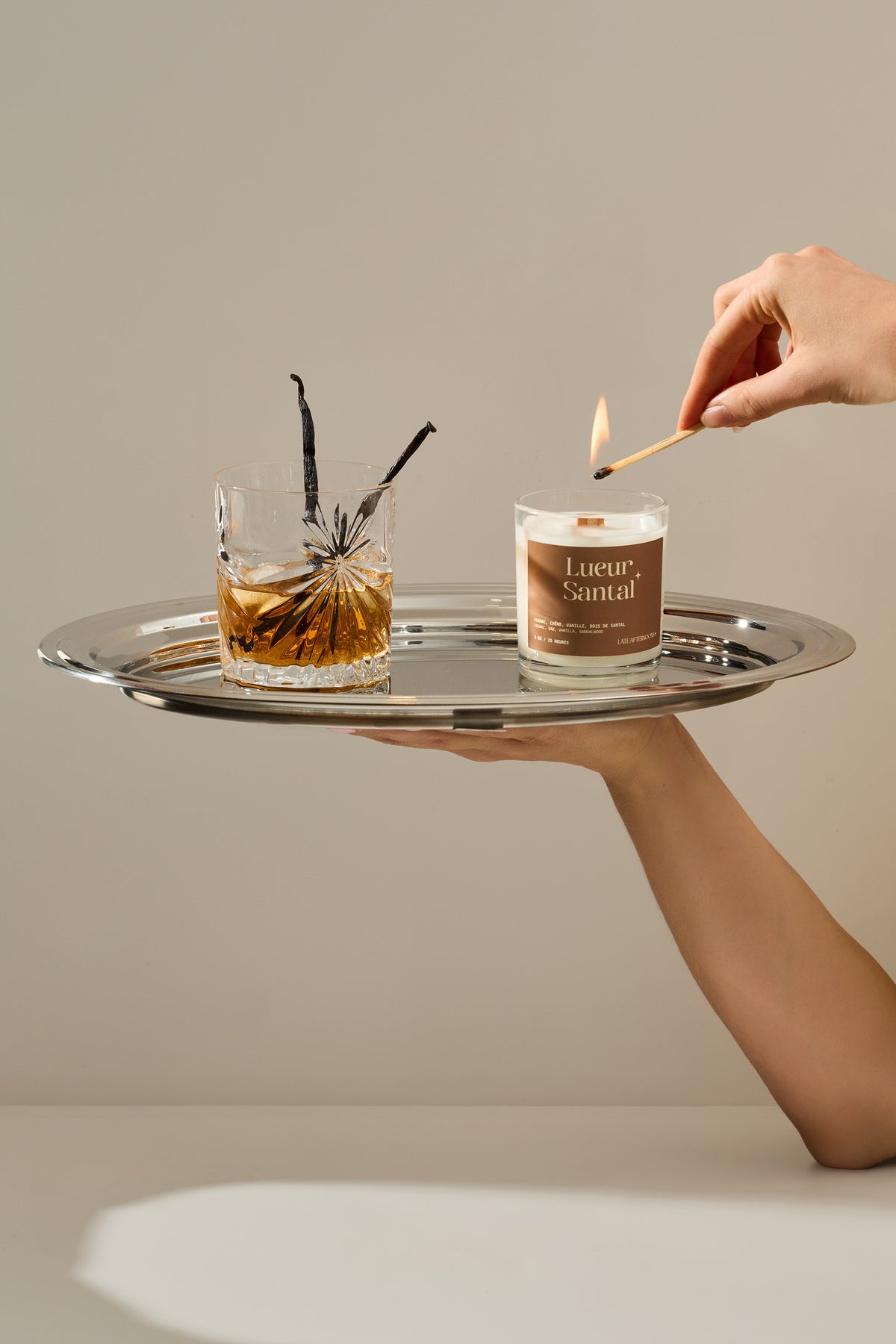 Une main tient un plateau d'argent et allume la bougie Lueur Santal avec une allumette, près d'un verre de whisky