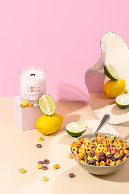 Bougie Sunday Funday allumée sur fond rose avec citrons, limes et un bol de céréales colorées